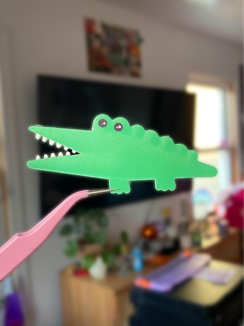 May include: A green paper alligator with a toothy grin and googly eyes is held up by a pair of pink tweezers.