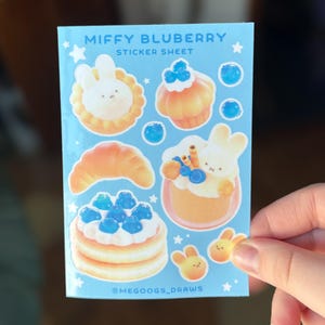 May include: A light blue sticker sheet featuring the text "MIFFY BLUBERRY STICKER SHEET". The sheet includes illustrations of baked goods and bunnies with blueberries. The artist's handle, @MEGOOGS_DRAWS, is also visible.