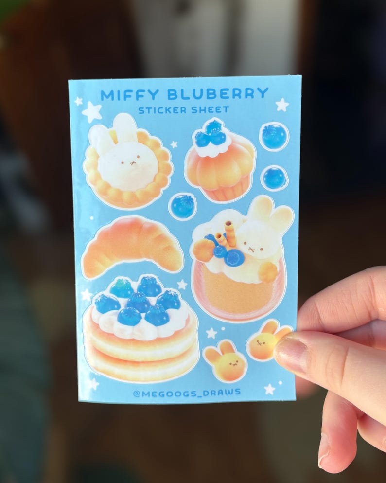 May include: A light blue sticker sheet featuring the text "MIFFY BLUBERRY STICKER SHEET". The sheet includes illustrations of baked goods and bunnies with blueberries. The artist's handle, @MEGOOGS_DRAWS, is also visible.