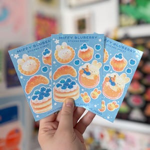 May include: Three light blue sticker sheets with the text "MIFFY BLUBERRY STICKER SHEET". Each sheet features illustrations of a bunny, pancakes, croissants, and other food items with blueberries. The stickers have a glittery effect.