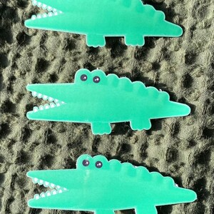 May include: Three green alligator stickers with white teeth and black eyes. The alligators are in a cartoon style and are facing to the right.
