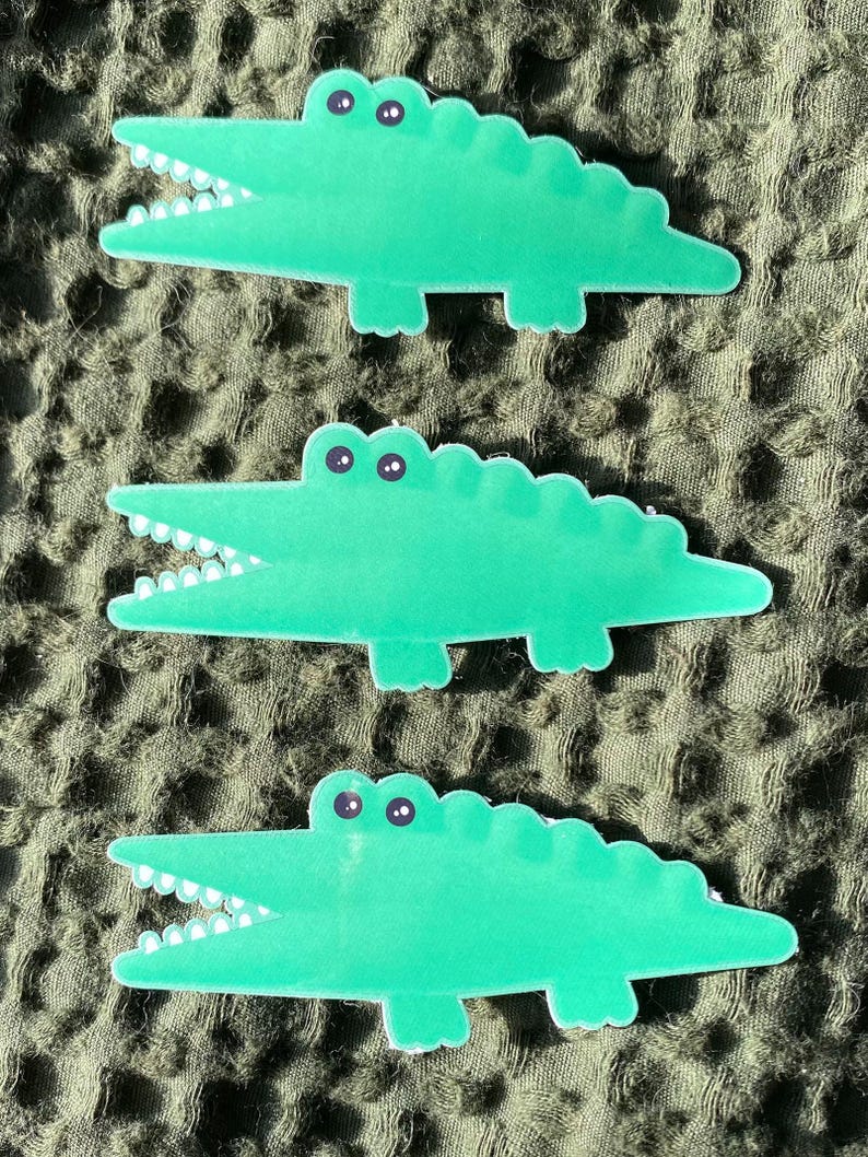 May include: Three green alligator stickers with white teeth and black eyes. The alligators are in a cartoon style and are facing to the right.