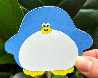 May include: A blue and white penguin-shaped paper cutout with yellow feet and a yellow beak. The penguin has a smiling face and is held in front of a green leafy background.