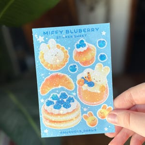 May include: A light blue sticker sheet featuring the text "Miffy Bluberry Sticker Sheet" and illustrations of desserts and bunnies. The sheet includes images of a croissant, pancakes, cupcakes, and other treats. The artist's name, @MEGOOGS_DRAWS, is at the bottom.