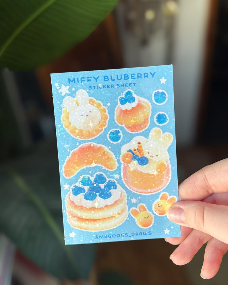 May include: A light blue sticker sheet featuring the text "Miffy Bluberry Sticker Sheet" and illustrations of desserts and bunnies. The sheet includes images of a croissant, pancakes, cupcakes, and other treats. The artist's name, @MEGOOGS_DRAWS, is at the bottom.