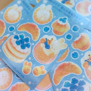 May include: Sheets of stickers featuring cute illustrations of bunnies, pancakes, croissants, and cupcakes with blueberries. The stickers have a light blue background with white stars and the text "Miffy Blueberry Sticker Sheet" and "@MEGOOGS_DRAWS".