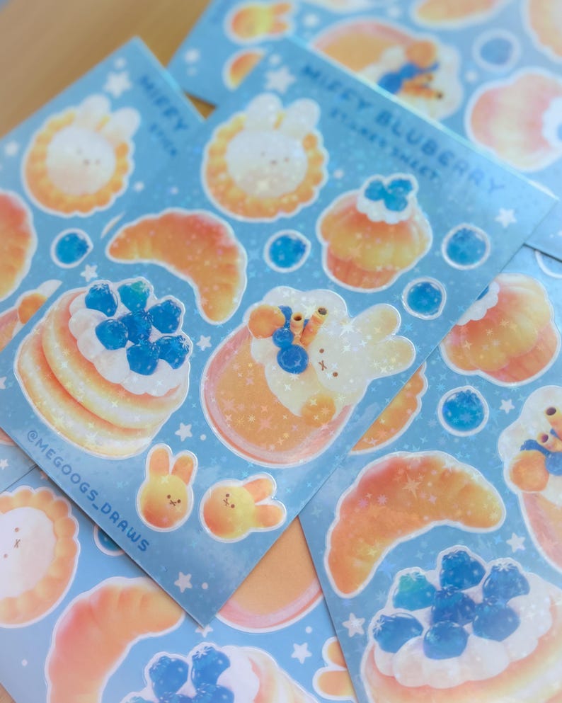 May include: Sheets of stickers featuring cute illustrations of bunnies, pancakes, croissants, and cupcakes with blueberries. The stickers have a light blue background with white stars and the text "Miffy Blueberry Sticker Sheet" and "@MEGOOGS_DRAWS".