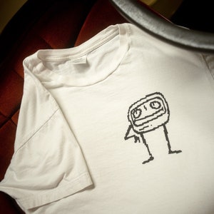 Peut inclure: T-shirt blanc avec un dessin pixelisé noir et blanc d'un personnage de dessin animé avec une grosse tête et un petit corps.