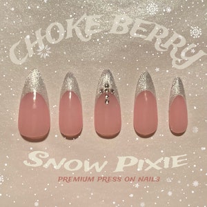 Puede incluir: Cinco uñas postizas de color rosa con un acento de purpurina plateada en la base. Las uñas tienen forma de óvalo largo y puntiagudo. El texto "CHOKE BERRY SNOW PIXIE PREMIUM PRESS ON NAILS" está impreso sobre un fondo blanco con un patrón de copos de nieve.