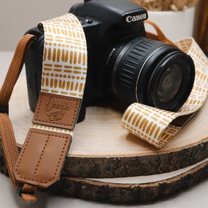 May include: A white and gold camera strap with a geometric pattern. The strap has a brown leather patch with the word "Dangle" embossed on it. The strap is attached to a black Canon camera.