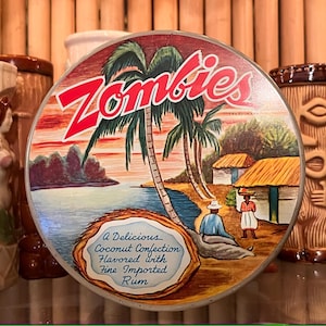 May include: A round tin container with a colorful illustration of palm trees, a beach, and two figures. The text "Zombies" is printed in red letters on the tin. The text "A Delicious Coconut Confection Flavored with Fine Imported Rum" is printed on the bottom of the tin.