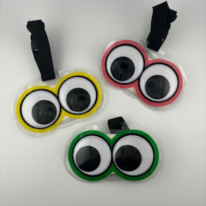 May include: Three novelty luggage tags with large cartoon eyes. One tag has yellow trim, another has pink trim, and the third has green trim. Each tag has a black strap and buckle for attachment.
