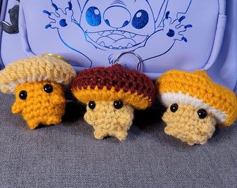 Può includere: Tre portachiavi a forma di fungo fatti a mano all'uncinetto. Ogni fungo ha un cappello di colore diverso: beige, marrone e giallo. I corpi dei funghi sono gialli. I portachiavi hanno occhi neri a bottone.