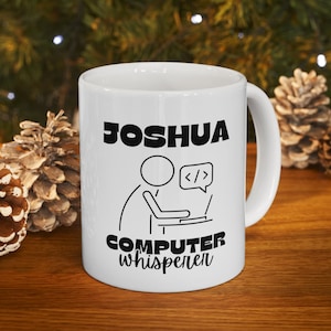 May include: White ceramic mug with a black design. The mug says "Joshua Computer Whisperer" with a stick figure of a person sitting at a computer with a speech bubble that says "</>"