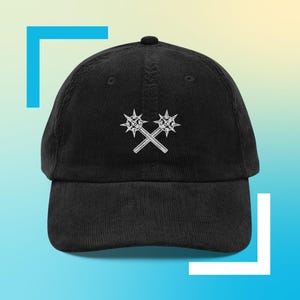 Puede incluir: Una gorra de béisbol de pana negra con un diseño bordado en blanco de dos mazas cruzadas con bolas con púas en los extremos.