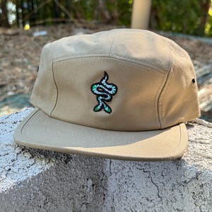 May include: A beige, five-panel cap with a flat peak. The cap has a green and white embroidered snake design on the front. The cap is made of a sturdy fabric and has a structured fit, suitable for casual wear.