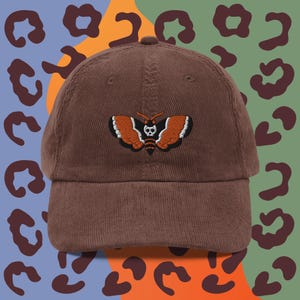 May include: A brown corduroy baseball cap with an embroidered black and orange moth with a skull design.