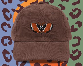 May include: A brown corduroy baseball cap with an embroidered black and orange moth with a skull design.