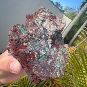 May include: A multi-coloured rock with an uneven surface. The rock features a mix of dark brown, deep red, and bright green patches. The texture is rough and irregular, indicating a natural mineral composition. The rock is held in a hand.