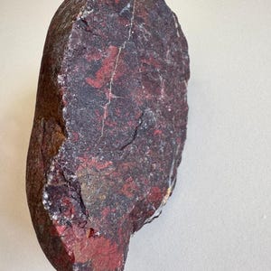 May include: A large, irregularly shaped rock with a dark maroon base colour and red and black veining. The rock has a rough, textured surface and a few visible cracks. The overall shape is elongated and slightly rounded.