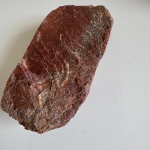 May include: A rough, reddish-brown stone with a textured surface. The stone has a rectangular shape with rounded edges and a mix of light and dark shades. The surface displays a network of fine lines and cracks.