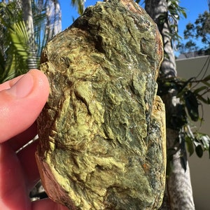 May include: A roughly textured rock displaying a blend of green, brown, and yellow tones. The natural stone has a rugged surface with visible ridges and colour variations. It is held in a hand against a backdrop of palm trees and a clear blue sky.