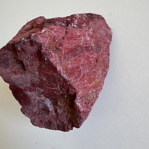 Puede incluir: Una roca tosca, de color rosa rojizo, con una superficie texturizada. La piedra muestra una mezcla de rojo intenso y tonos rosados más claros, con vetas minerales visibles. La forma irregular y las variaciones de color sugieren un espécimen natural sin pulir.