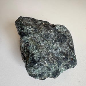 May include: A dark green rock with a rough, textured surface. The stone displays a mix of dark and light green shades, with some areas appearing nearly black. The rock is irregularly shaped, suggesting it is a natural mineral specimen.