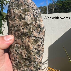 Könnte beinhalten: Ein großer, unregelmäßig geformter Stein mit nassem Aussehen. Der Stein weist eine Mischung aus rosa, grünen und schwarzen gesprenkelten Mustern auf. Der Text "Wet with water" ist auf der rechten Seite des Bildes zu sehen. Der Stein wird von einer Hand gehalten.