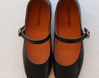 May include: A pair of black leather shoes with a buckle strap. The shoes have an orange insole and the text "MIH Studio" is printed on the inside of each shoe.