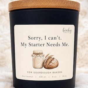 Op de afbeelding: Zwarte kaars met een houten deksel. Het etiket luidt "Sorry, I can't. My Starter Needs Me." met illustraties van zuurdesem en brood. Het is een soja-kokoskaars, 230 ml, met een brandtijd van 35+ uur.