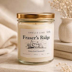 Op de afbeelding: Een witte kaars in een glazen pot met een gouden deksel. Het etiket vermeldt: SMELLS LIKE Fraser's Ridge, Dinna Fash Sassenach!, Soy Coconut | 225 g | 8oz | 35 h+. Het etiket bevat een zwart-witte lijntekening van een hut in de bergen.