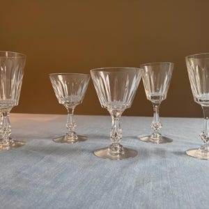 Pode incluir: Cinco taças de vinho de cristal transparente de tamanhos variados são dispostas sobre uma superfície azul clara. Cada taça apresenta uma haste decorativa e uma taça texturizada e facetada. As taças estão vazias e prontas para uso.
