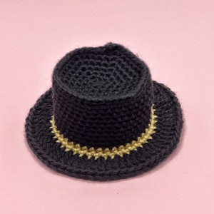 May include: A miniature black crochet top hat with a gold-colored band around the base of the crown. The hat is set against a soft pink background, showcasing the handcrafted details.