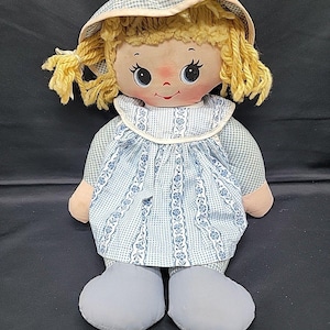 May include: A vintage rag doll with a painted face, yellow yarn hair, and a blue and white checkered bonnet and dress. The doll has light blue feet and a pinkish-tan skin tone. The dress features a floral pattern.