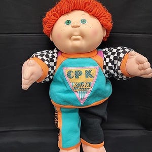 May include: A vintage Cabbage Patch Kids doll with red yarn hair, green eyes, and freckles. The doll wears a teal, orange, and black outfit with the text "CPK Kids". The sleeves have a black and white checkered pattern.