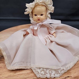 Op de afbeelding: Een vintage babypop met een roze jurk en een witte kanten muts. De pop zit op lichtroze stof met een kanten rand. Het gezicht van de pop heeft geschilderde kenmerken, waaronder bruine ogen en rode lippen. De achtergrond is donker.