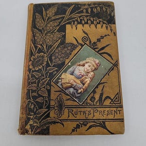 Könnte beinhalten: Ein gebundenes Vintage-Buch mit dem Titel Ruth's Present mit einem beigefarbenen und schwarzen Einband, einem geprägten Blumenmuster und einer zentralen rautenförmigen Illustration einer Person, die eine Puppe hält. Das Design enthält schwarze Details.