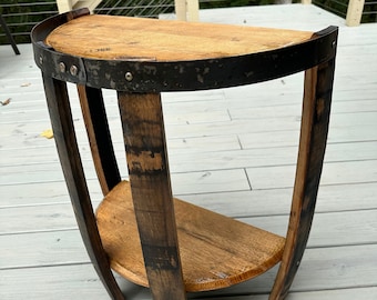 May include: A half-circle wooden side table made from reclaimed barrel wood. The table features a dark metal band around the top and a lower shelf. The legs are curved and made from the same barrel wood. The table is approximately 24 inches tall.