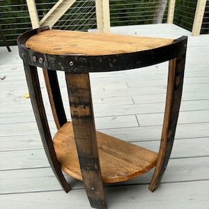 May include: A half-circle wooden side table made from reclaimed barrel wood. The table features a dark metal band around the top and a lower shelf. The legs are curved and made from the same barrel wood. The table is approximately 61 cm tall.
