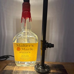 May include: A unique lamp made from a Maker's Mark whiskey bottle, featuring a red wax seal and a black pipe frame. The lamp sits on a wooden base and has a black cord.