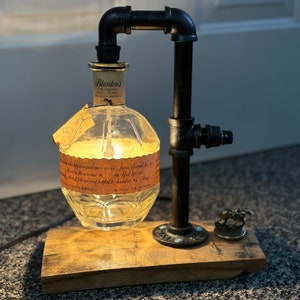 May include: A unique lamp made from a repurposed Blanton's bourbon whiskey bottle. The clear glass bottle is mounted on a wooden base and connected to black metal pipes. A small horse and jockey figurine sits on the base.
