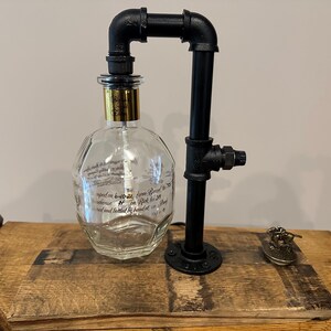 May include: A unique lamp made from a repurposed liquor bottle and black metal pipes. The clear glass bottle has gold accents and is mounted on a wooden surface. A small decorative object sits nearby.