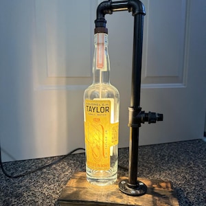 May include: A unique lamp made from a repurposed whiskey bottle, featuring a black pipe frame and a wooden base. The bottle has a yellow label with the text "COLONEL E.H. TAYLOR SMALL BATCH". The lamp is a creative blend of industrial and rustic design.