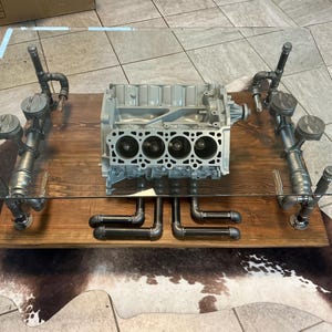 May include: A unique coffee table featuring a glass top supported by an engine block and metal piping. The table has a rectangular wooden base and is designed with industrial elements. The engine block is silver and has four cylinders.