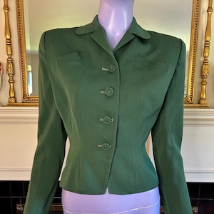 May include: A forest green long-sleeved blazer featuring a notched collar, structured shoulders, and a front button closure with four matching green buttons. The garment includes two decorative horizontal flap details on the upper chest area.