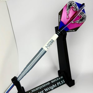 May include: A silver dart with a black and white "Nitro" logo on the barrel. The dart has a pink and blue flight with a black and white scorpion design. The dart is in a black stand with the text "NIGHTQUEEN3D - REPLICA #1 Peter Wright PL15"