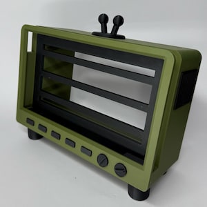 May include: An olive green and black decorative object, reminiscent of a retro television set. It has a rectangular frame with black horizontal slats, black knobs, and a pair of black antenna-like elements on top. The design is minimalist and the object is olive green.