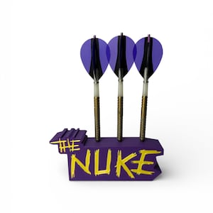 May include: Three darts with purple and black flights are displayed in a purple holder. The holder has the words "THE NUKE" in yellow. The darts have brass-coloured shafts and white and black accents.