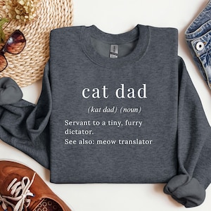 Może przedstawiać: Ciemnoszara bluza z białym napisem "cat dad" i definicją. Definicja brzmi: "Sługa małego, futrzanego dyktatora. Zobacz też: tłumacz miau."